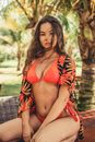 Young alluring woman in bright trendy swimsuit sitting against tropical trees and looking at camera