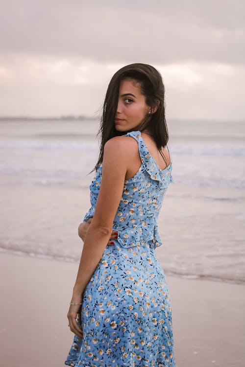 Back view of young gentle female tourist in sundress with hair on face looking at camera over shoulder on ocean coast