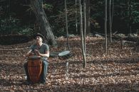 Full body of concentrated middle aged male musician in casual clothes and hat playing conga percussion instrument sitting in autumn park with fallen leaves near drum