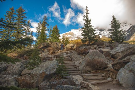Experience the beauty of Banff with a hiker on a rocky trail surrounded by trees and snow-capped mountains.