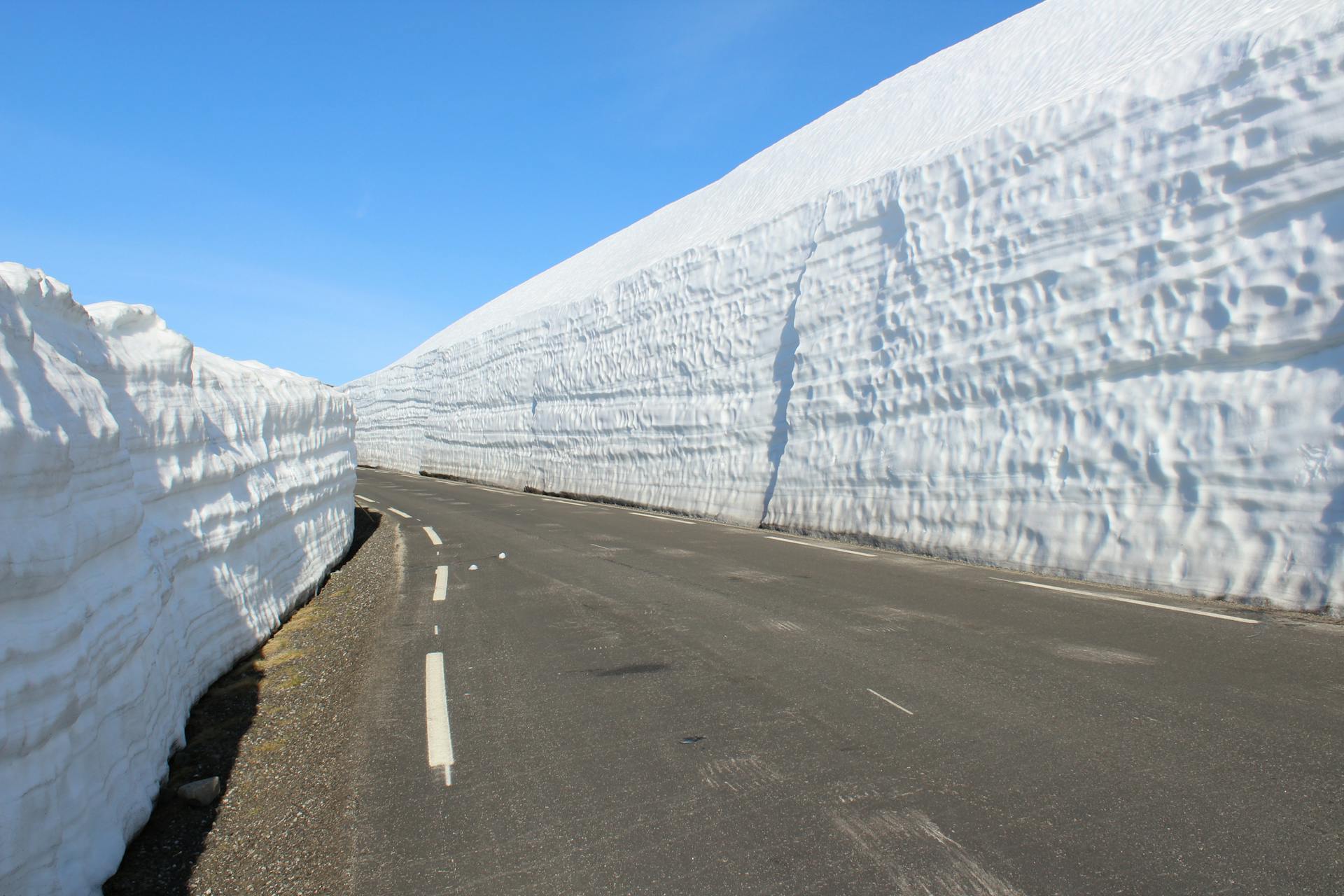 Road lined with tall snow walls under a clear blue sky, showcasing winter's beauty.