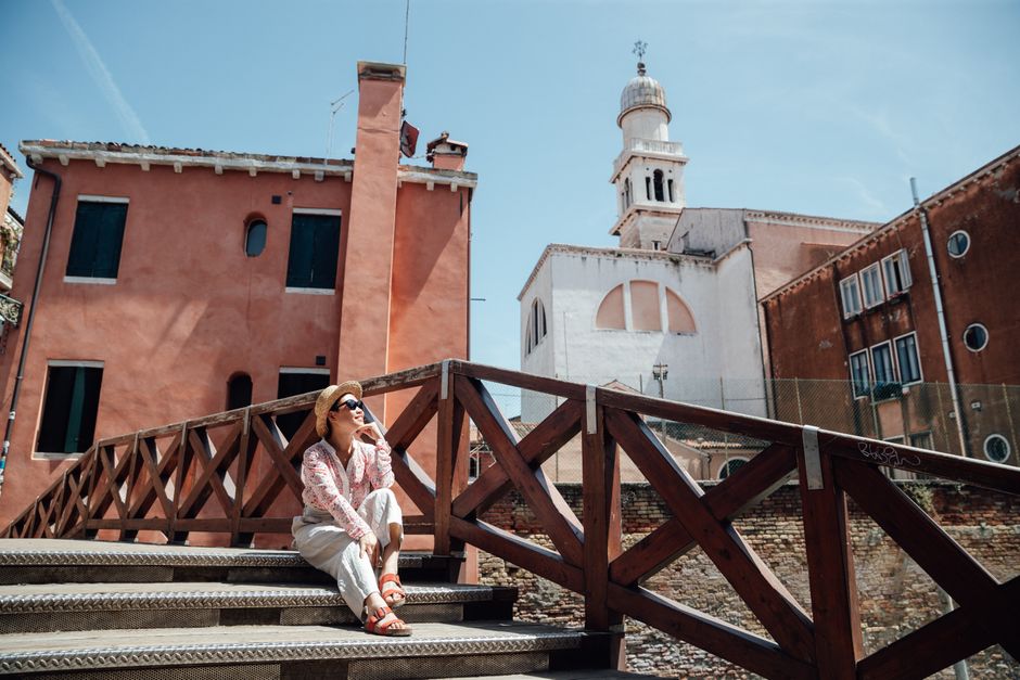 Full body of female traveler in hat and sunglasses sitting on stairs at wooden railing near aged residential houses with towers during summer vacation