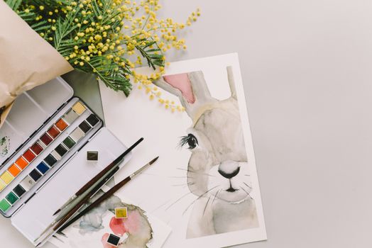 Watercolor painting of a bunny surrounded by mimosa flowers and art supplies.