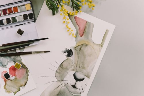 Free Artistic watercolor painting of an Easter bunny with paintbrushes, watercolors, and mimosa flowers. Stock Photo