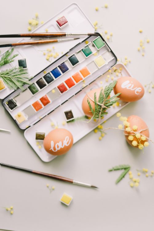 Free Colorful Easter eggs painted with watercolors, featuring yellow mimosas and art supplies in a flat lay design. Stock Photo