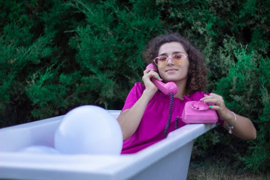 A woman sits in a bathtub outdoors talking on a vintage pink rotary phone, wearing sunglasses.