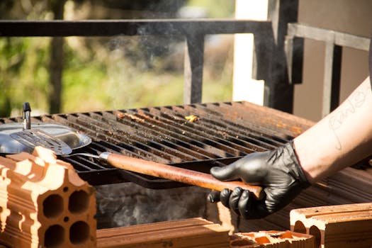 Outdoor BBQ scene with a gloved hand holding a spatula over a grill.