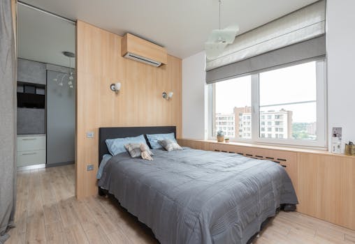 Interior of light modern apartment with bed near window and jalousie with windowsill with decorative elements near air conditioner and entrance to kitchen