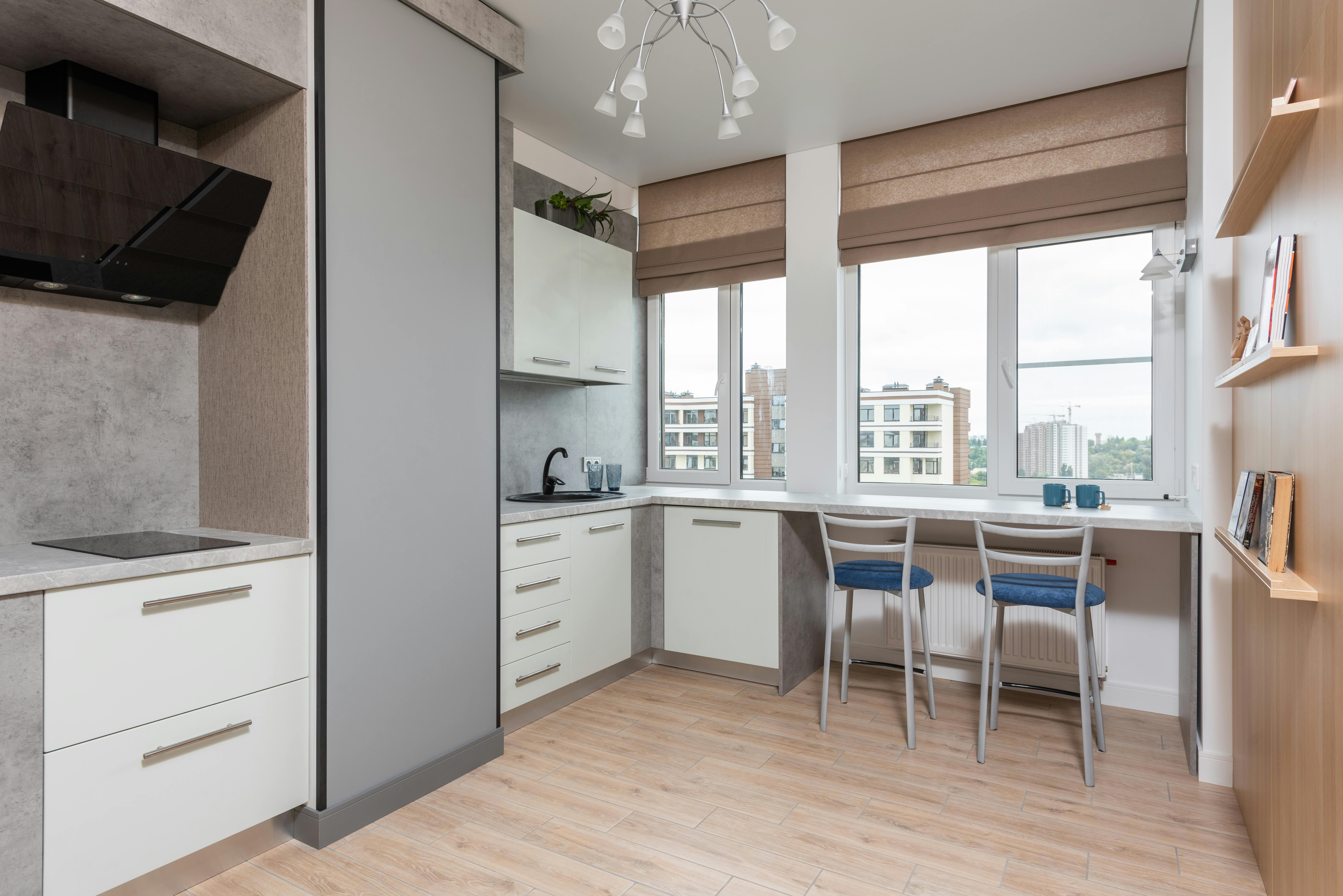 Interior of light kitchen with white cabinets and cupboards with sink and tap near counter with chairs near window with jalousie near shelves with books