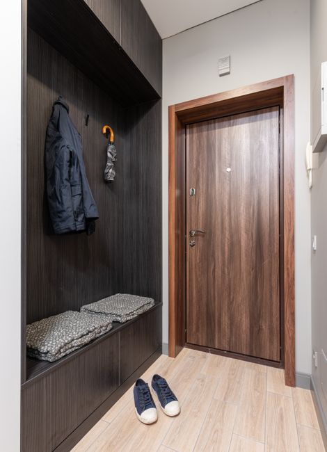 Modern apartment entryway with a wooden wardrobe and white shelving, organized with jackets, umbrella, and sneakers for easy access.