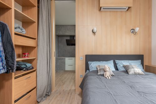 Free Modern room interior with bed with pillows and soft rabbit toy near wardrobe with shelves and clothes near entrance to kitchen with curtains in light apartment Stock Photo