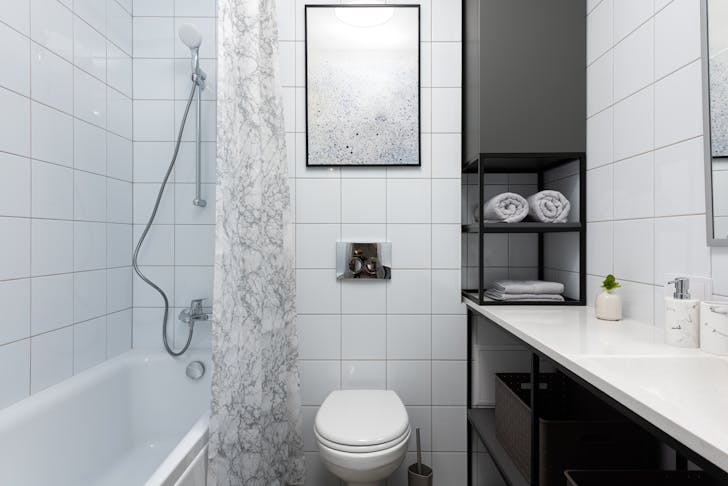 Interior of light bathroom with toilet near bathtub with shower and curtain near counter with shelves with towels and containers and decorative objects near white tile