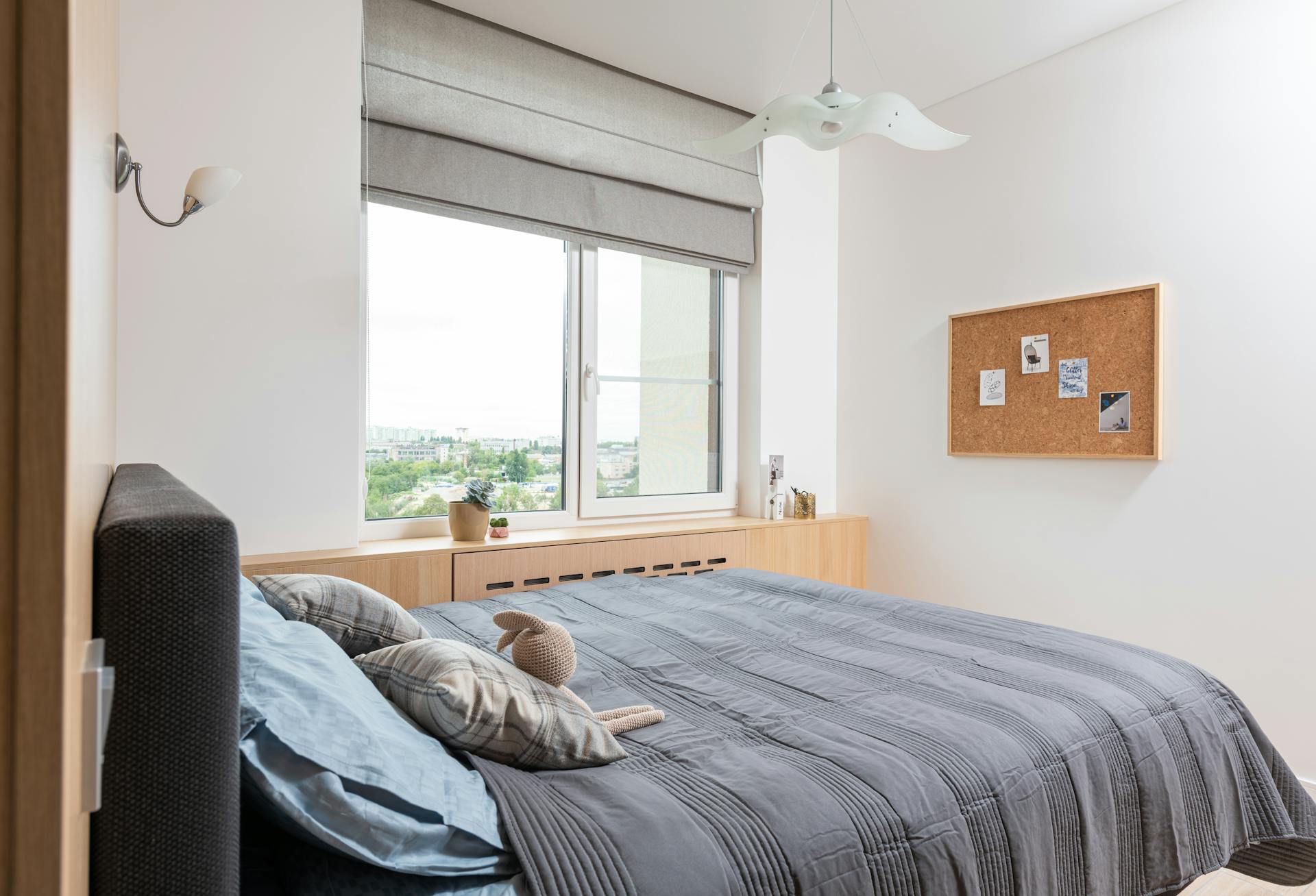 Interior of contemporary apartment with bed with pillows and soft bunny toy near window with windowsill with decorative elements with jalousie near lamps and wooden board on wall with photos