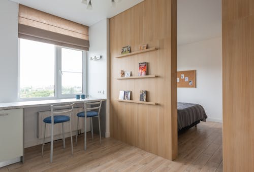 Interior of light flat with counter near chairs and windows with jalousie near shelves with books and entrance in room with bed in daylight