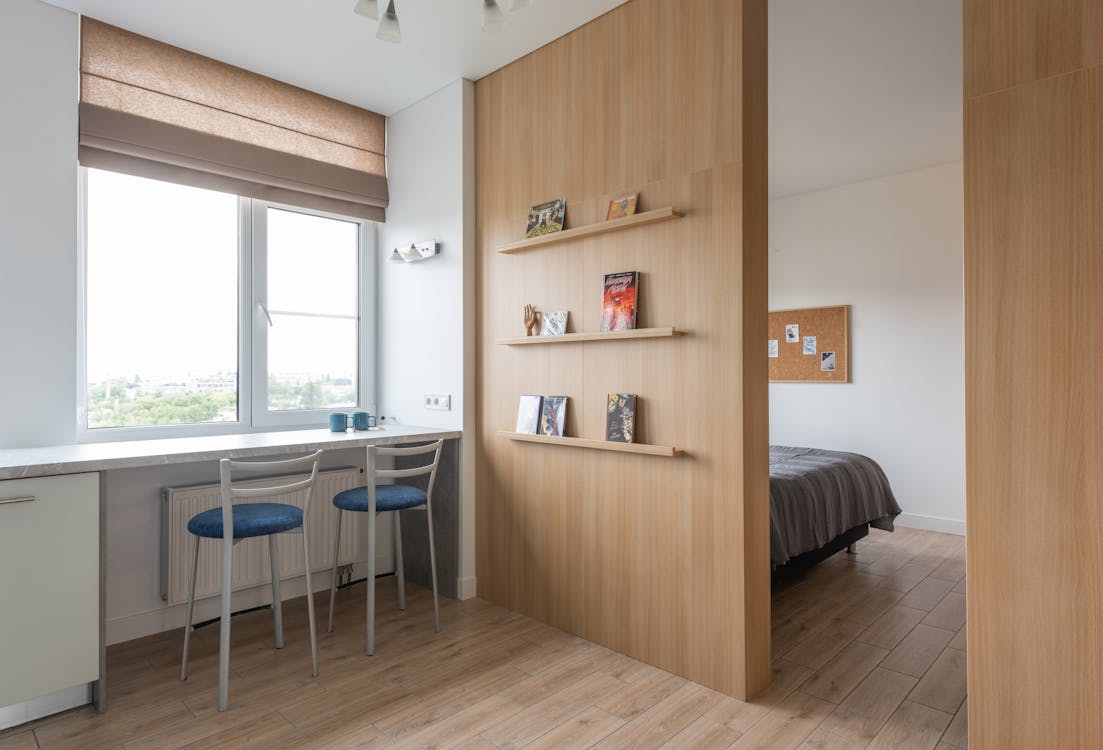 Free Interior of light flat with counter near chairs and windows with jalousie near shelves with books and entrance in room with bed in daylight Stock Photo