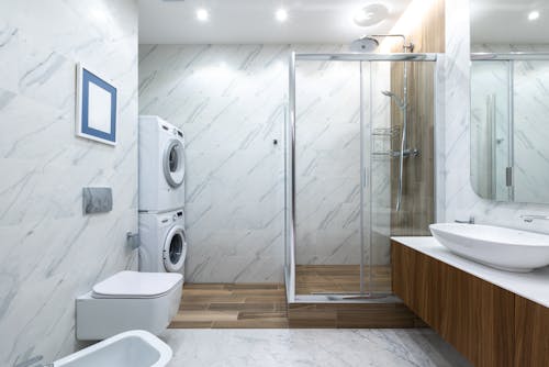 Contemporary bathroom toilet near tiled wall against cabinet with sink placed near shower cabin with glass wall and washing machines