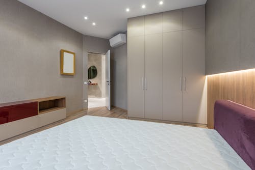 Interior of modern bedroom with white mattress on bed placed against cabinet near big closet and opened door in spacious apartment