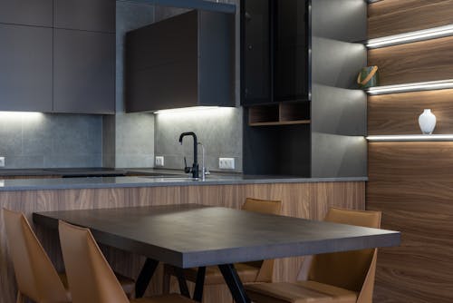 Contemporary black cupboards with sink in stylish kitchen near wooden table and chairs placed near wall with light decoration at home