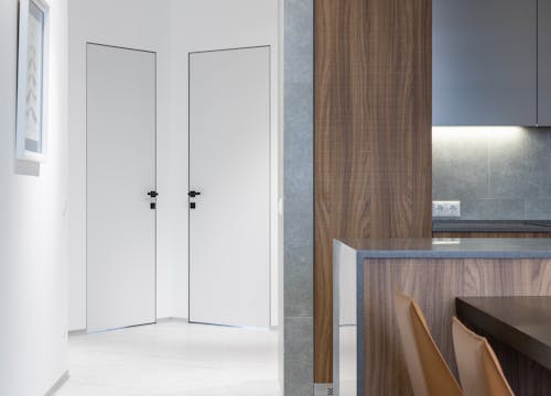 Hallway with white doors and walls near wooden kitchen counter with cupboards near table and chairs in modern apartment at home