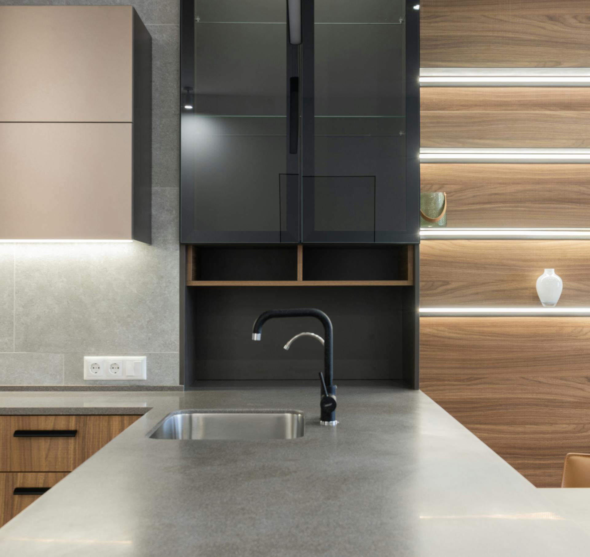 Gray kitchen counter with faucet and sink near cabinets and light decorations on wooden wall in modern apartment at home