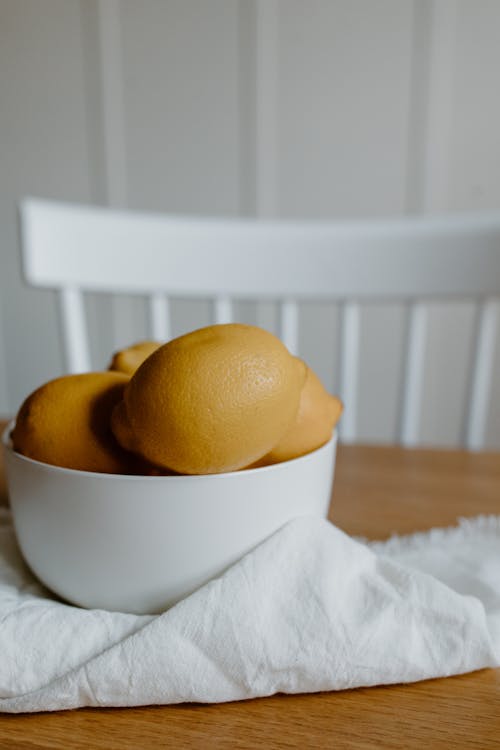 White bowl filled with yellow lemons on textile on wooden table near chair in light room
