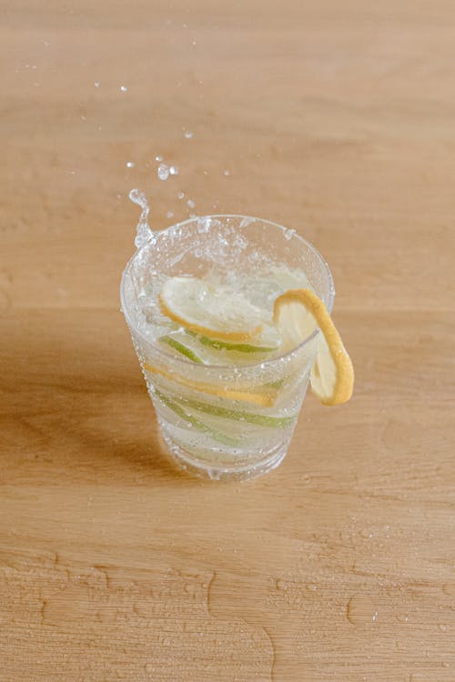 High angle of glass with transparent cocktail decorated with slices of limes and lemons placed on wooden floor in light room