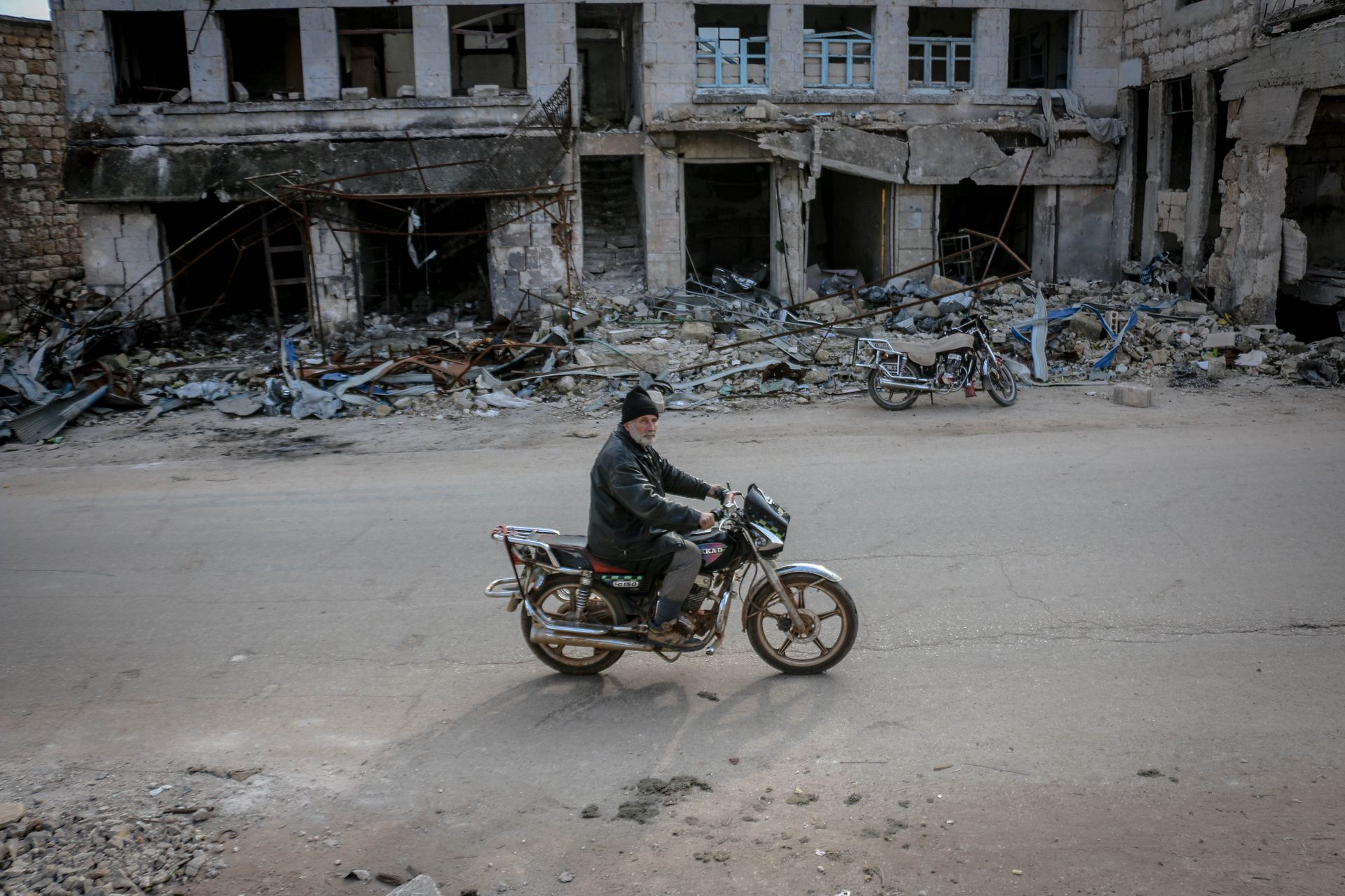 A man rides his motorbike past ruins in Idlib, Syria, showcasing post-war decay.