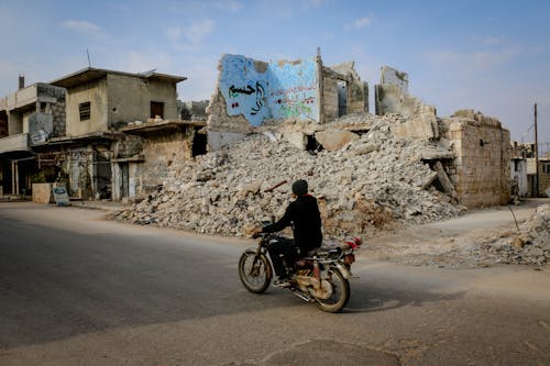 Free Full body side view of anonymous man riding motorcycle on road near heap of ruins and destroyed buildings in poor district Stock Photo
