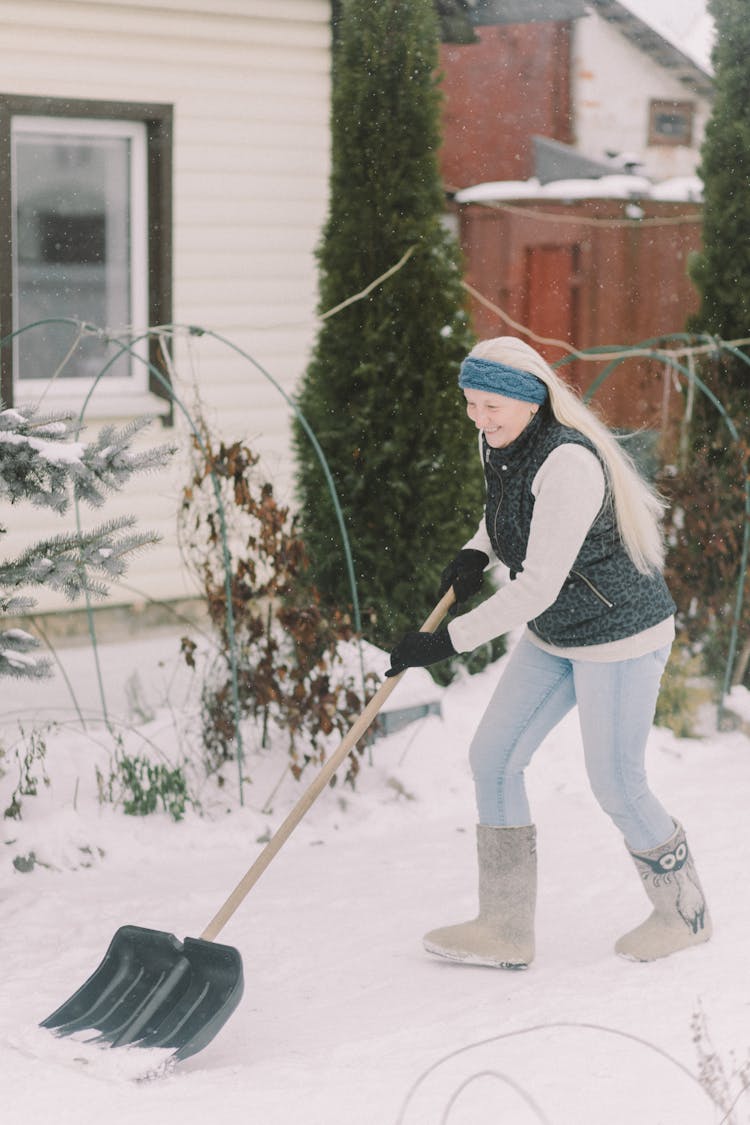 Woman In Long Sleeve Shirt And Denim Pants Plowing Snow With Shovel