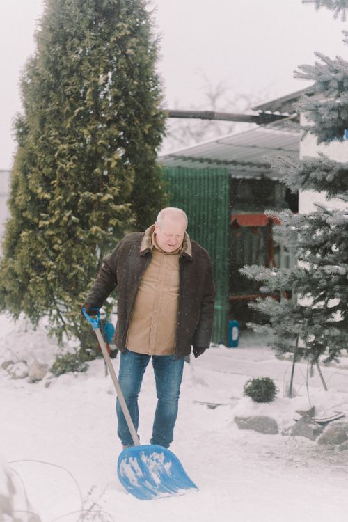 Free Elderly man in a brown jacket shoveling snow outdoors in winter season. Stock Photo