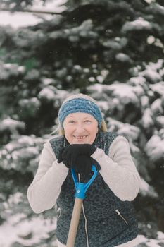 Smiling elderly woman in winter attire holding a shovel outdoors in snowy scenery.