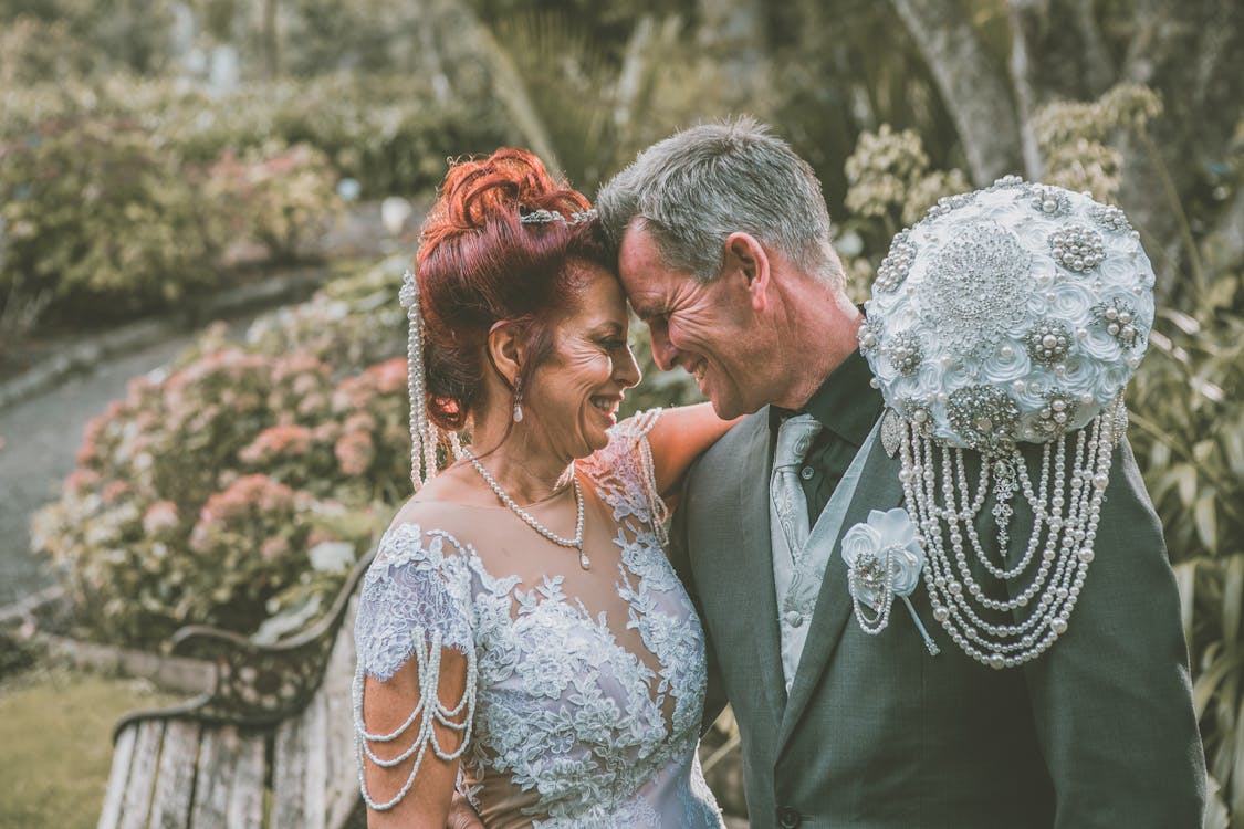 Free Side view of happy middle aged bride and groom in elegant outfits smiling and looking at each other while touching foreheads in green park on wedding day Stock Photo
