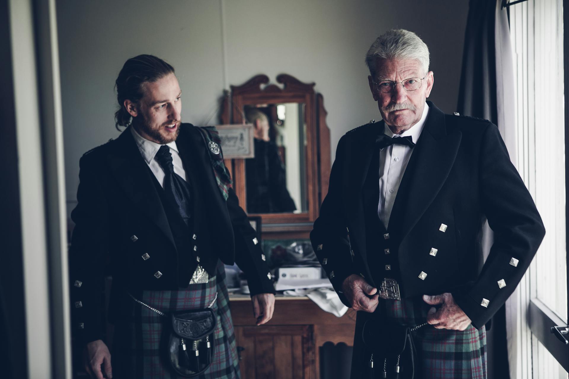 Focused young and senior Scottish men wearing Highland traditional dress with kilt jacket and sporran while standing together in classic styled room