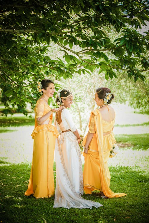Free Full body cheerful young Sinhalese bride in elegant white dress laughing while standing on lawn in green park with happy best friends in similar outfits on wedding day Stock Photo