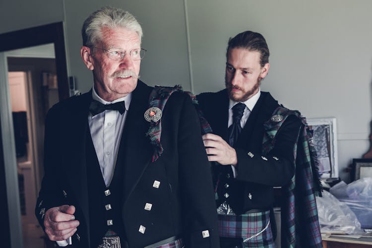 Young Man With Elderly Father Wearing Traditional Highland Dress In Room