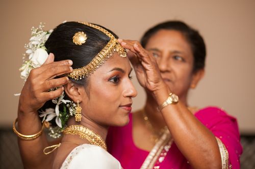 Free Mature ethnic woman in bright sari putting traditional golden Kandyan jewelry on head of elegant Sinhalese bride before wedding ceremony Stock Photo
