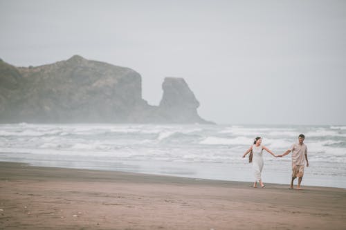 Joyful young ethnic couple holding hands and walking on sandy seashore against overcast sky