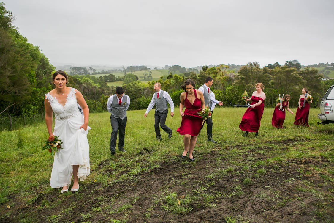 Free Full body of young bride in white dress with flower bouquet in hand walking on grassy meadow with bridesmaids and groomsmen before outdoor wedding ceremony Stock Photo