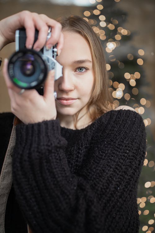 Free Attractive positive female in knitted black sweater looking at camera and taking pictures on professional photo camera while standing against shining garland Stock Photo