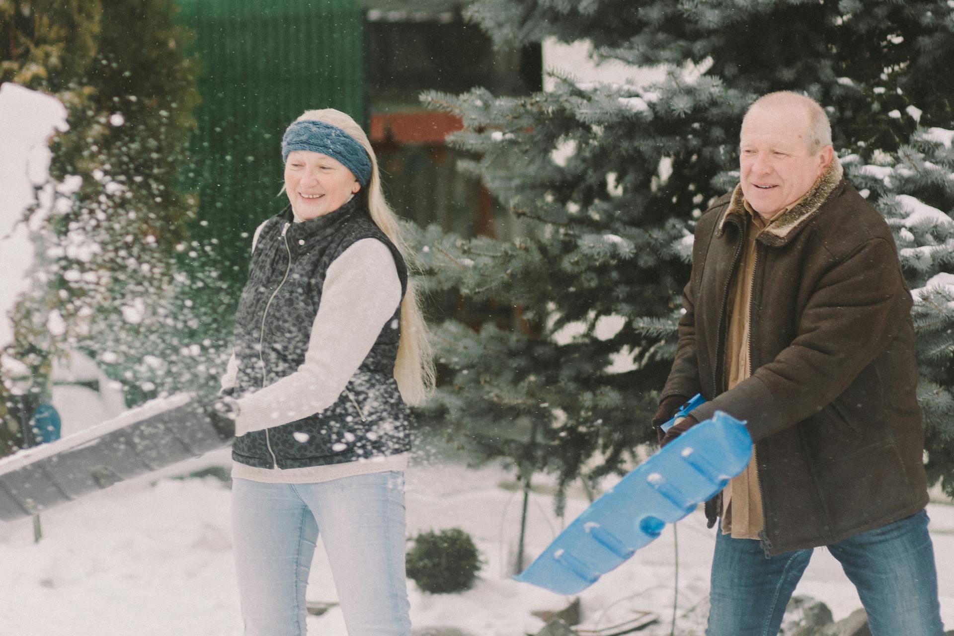 An elderly couple happily shovels snow together outdoors, embracing the winter season.