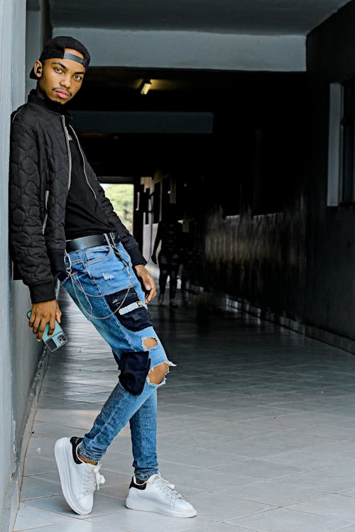 Man Wearing Black Jacket and Ripped Denim Jeans Leaning on Wall