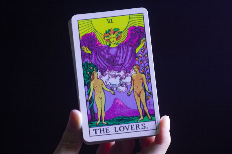 A hand holding The Lovers tarot card against a dark background