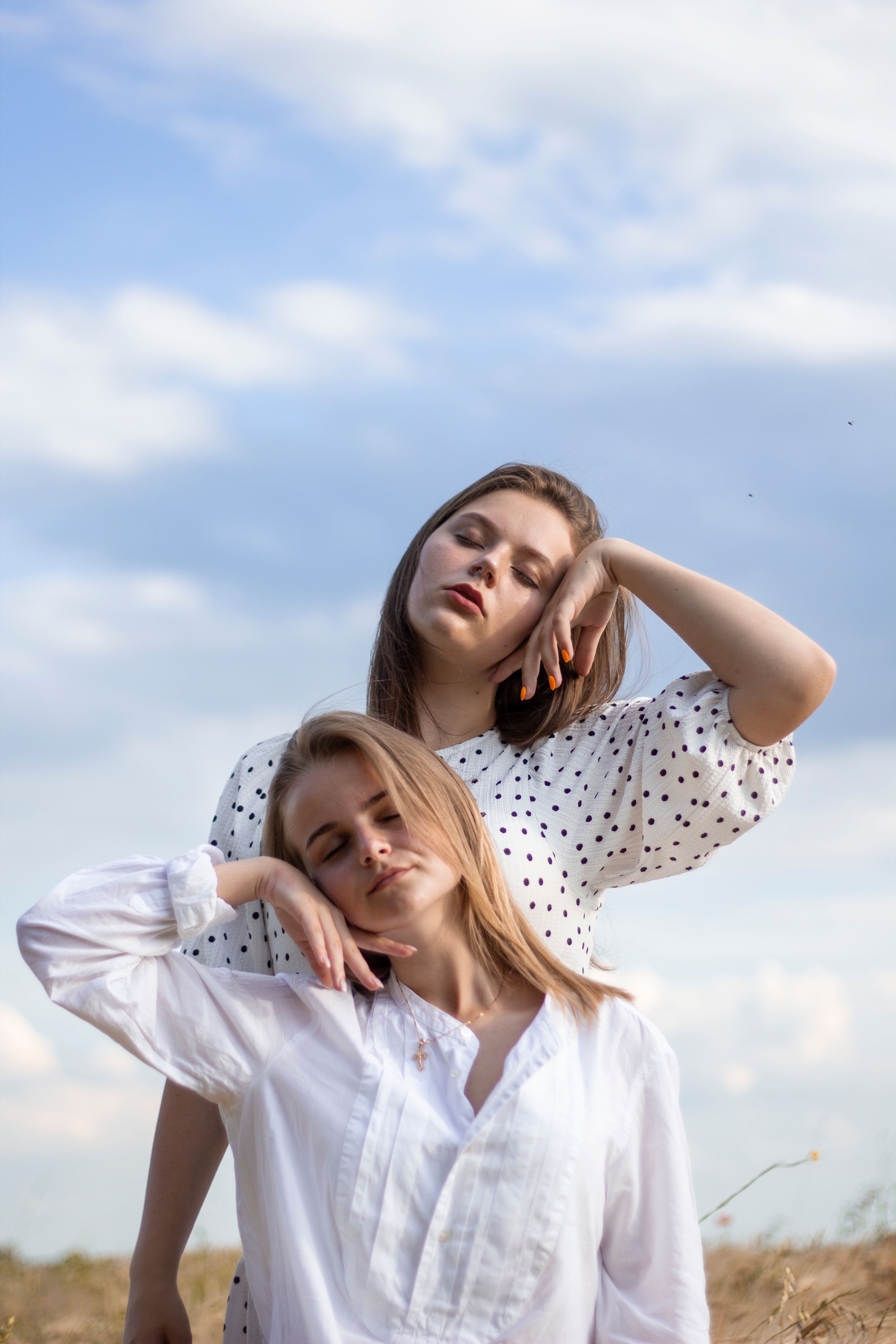Two women in white tops pose with serenity under a blue sky and clouds.