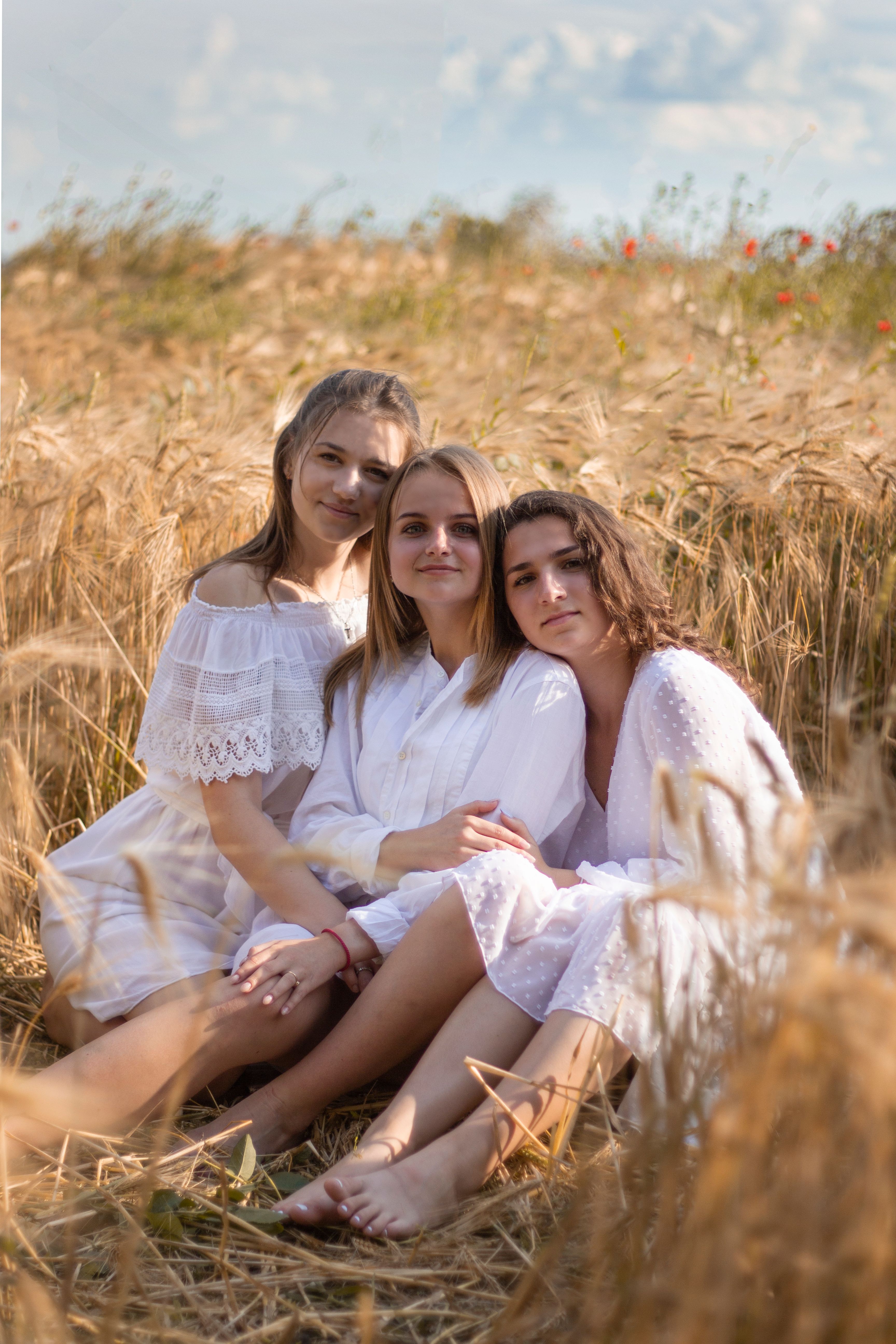 Three young women sitting together in a sunny wheat field, enjoying the outdoors and smiling.
