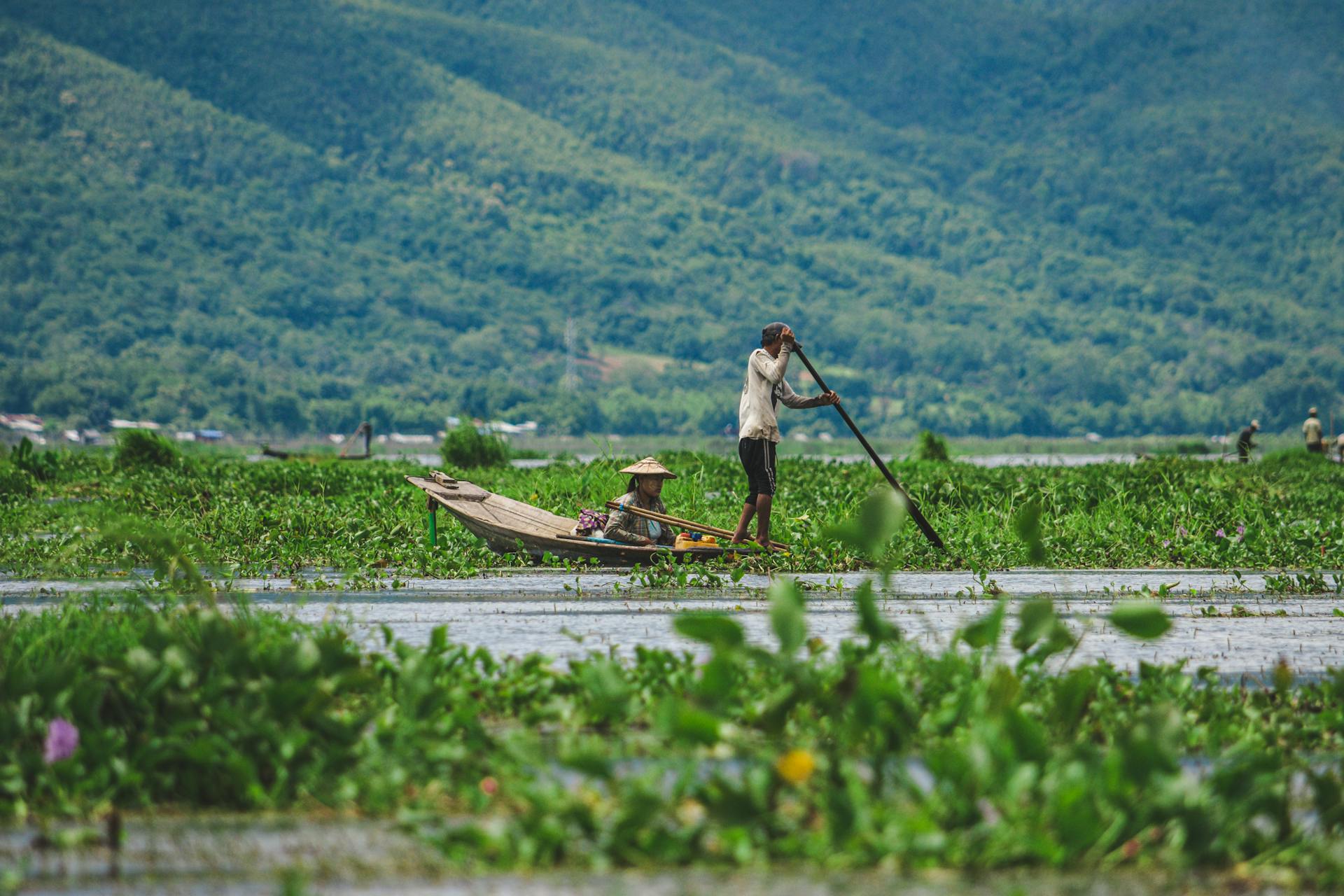 A man rows a wooden boat with a child on Inle Lake surrounded by lush greenery.