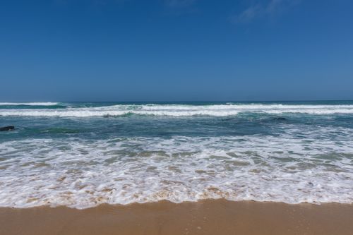 Free Calm ocean waves crashing onto a sandy beach under a clear blue sky, perfect for nature lovers. Stock Photo