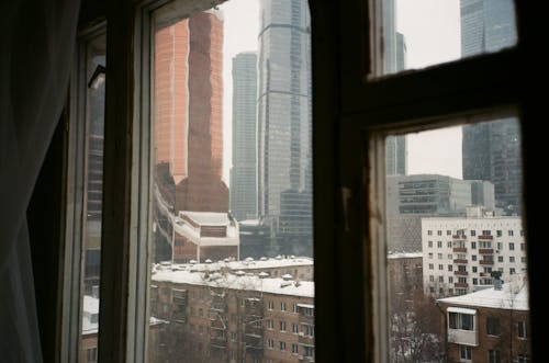 Through apartment window view of modern urban city architecture with snowy residential buildings and skyscrapers on overcast winter day