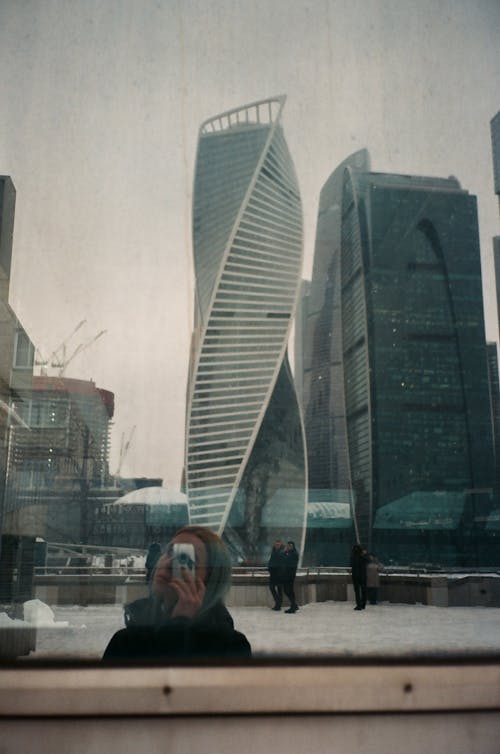 Female in outerwear taking photo on photo camera of modern Moscow City skyscrapers reflecting on glass wall on winter day