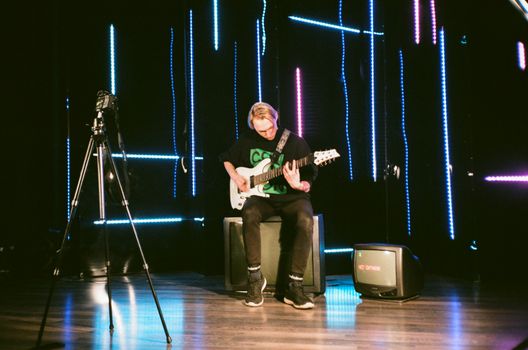 Full body focused young professional male musician playing bass guitar while sitting on floor speaker on stage during rehearsal