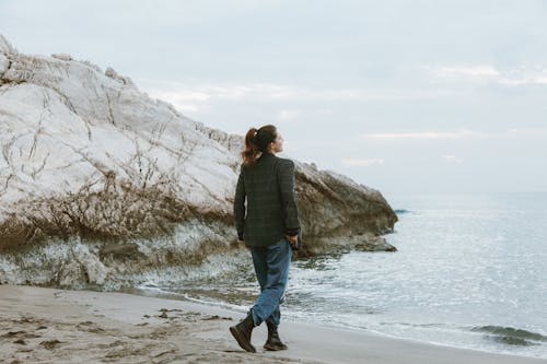 Woman Wearing a Jacket, Denim Jeans and Boots Walking on the Seashore